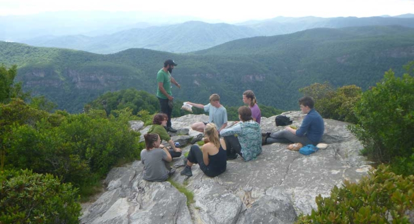 A group of students rest on a rocky overlook, high above the vast blue ridge mountains below. 